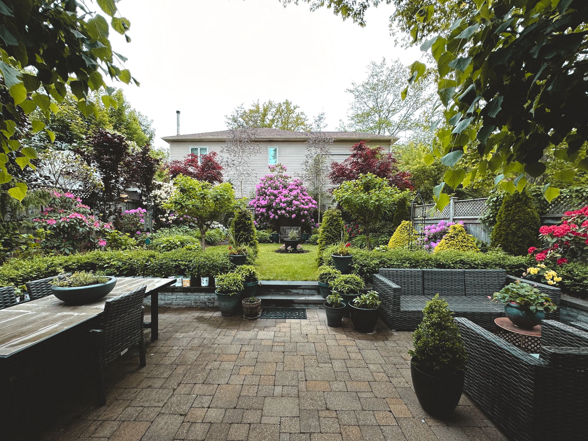 A paver patio framed by a lush, flowering backyard garden and manicured lawn